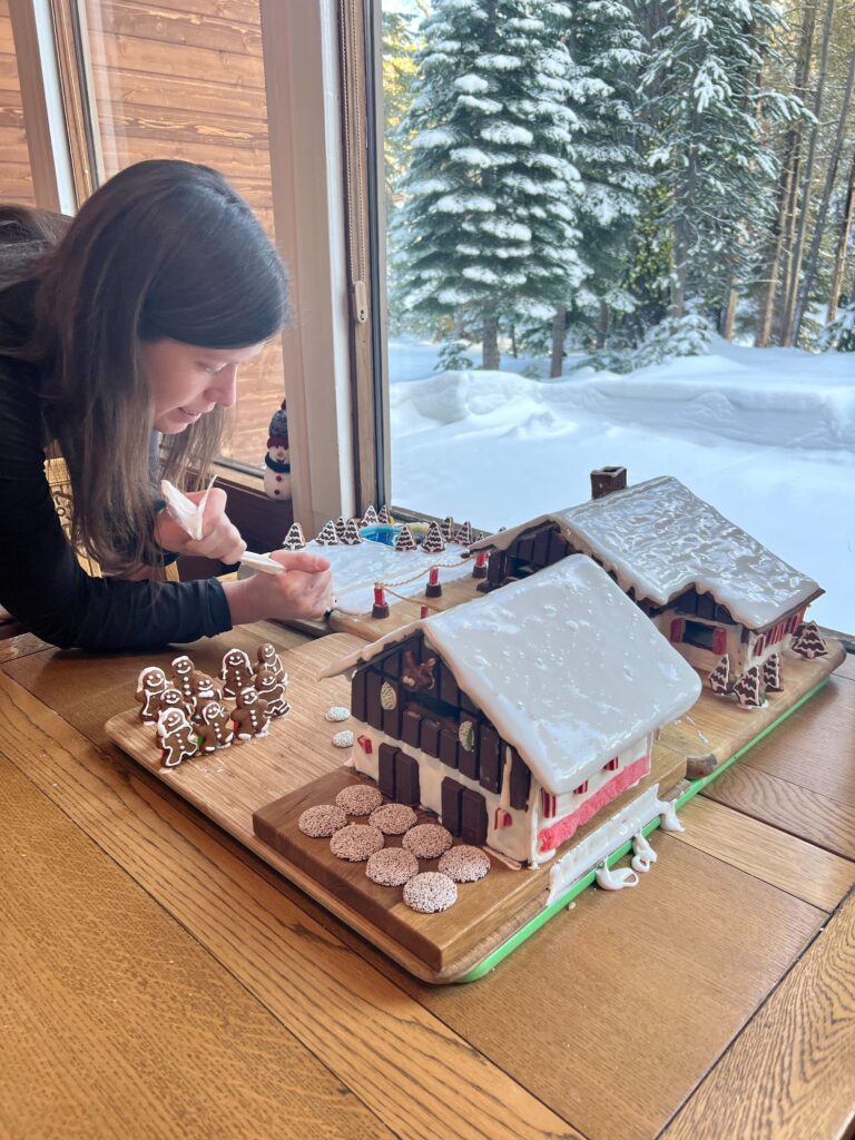 Kate Hake decorates a gingerbread house village on a wooden table by a window, with snow-covered trees and ground visible outside.