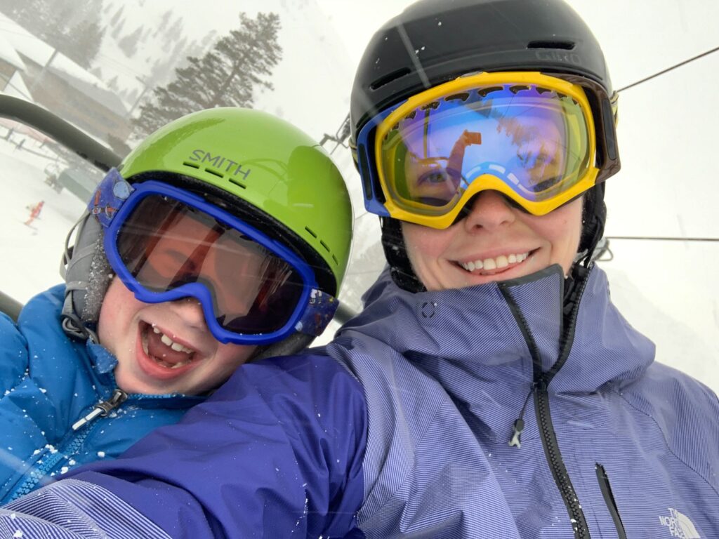 Two of our people, dressed in ski gear including helmets and goggles, smile whilst riding a ski lift on a snowy day. Snow-covered trees and slopes are visible in the background.