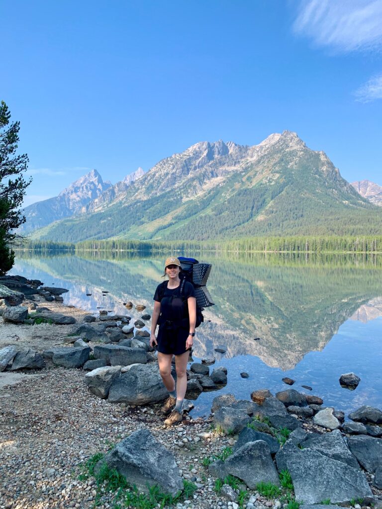 Kate Hake, wearing a rucksack, stands by a serene lake with mountains and trees reflected in the calm water under a clear blue sky.