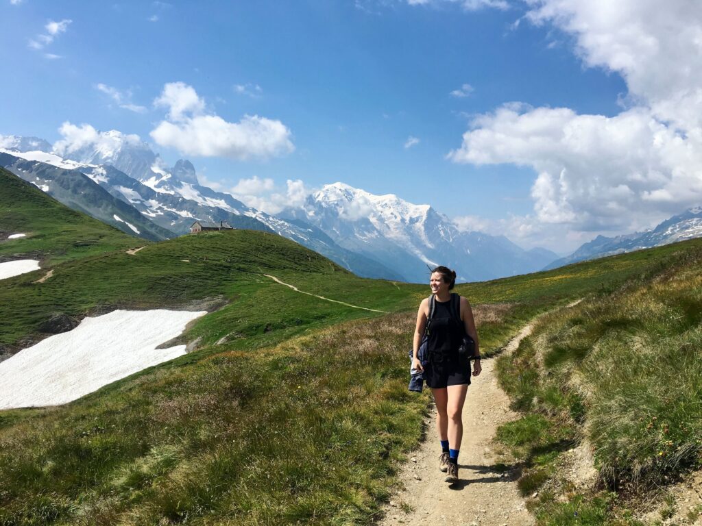 Kate Hake hikes along a narrow mountain trail with green hills, patches of snow, and snow-capped peaks in the background under a partly cloudy sky.