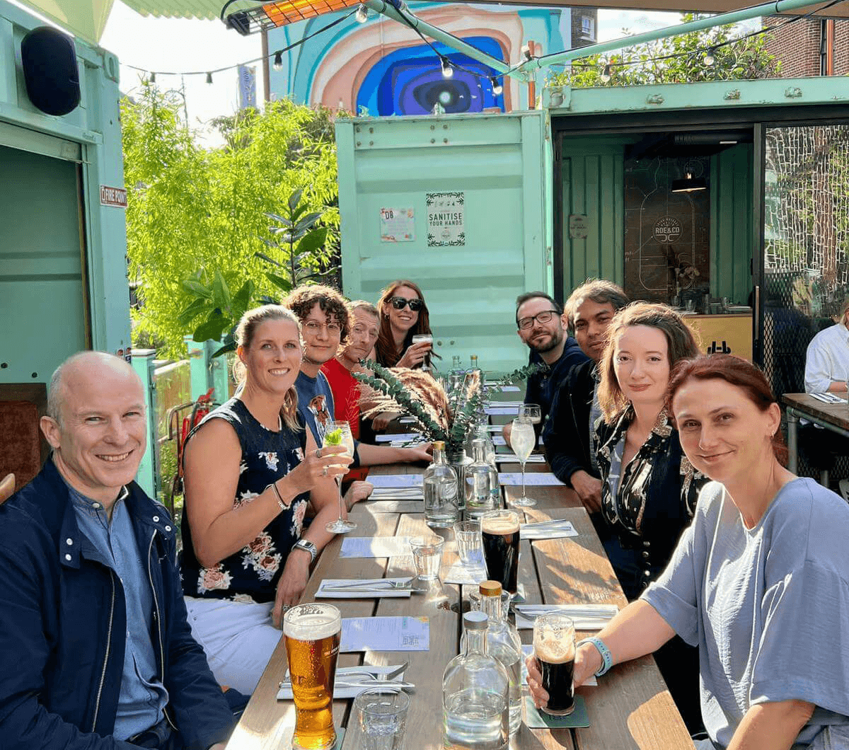 A group of ten people sit around an outdoor restaurant table, enjoying drinks and smiling. Sunlight creates a cheerful atmosphere with plants and colourful mural-painted containers in the background.