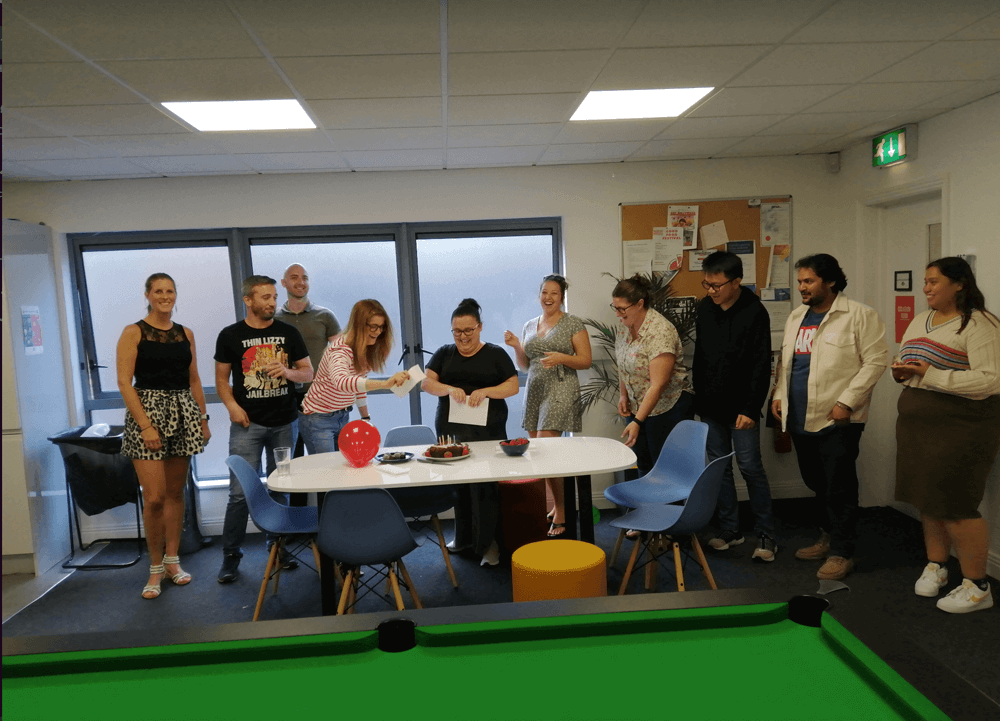 A group of people stand around a table with balloons, cake, and papers in a brightly lit room, smiling and talking, as a green snooker table is visible in the foreground.