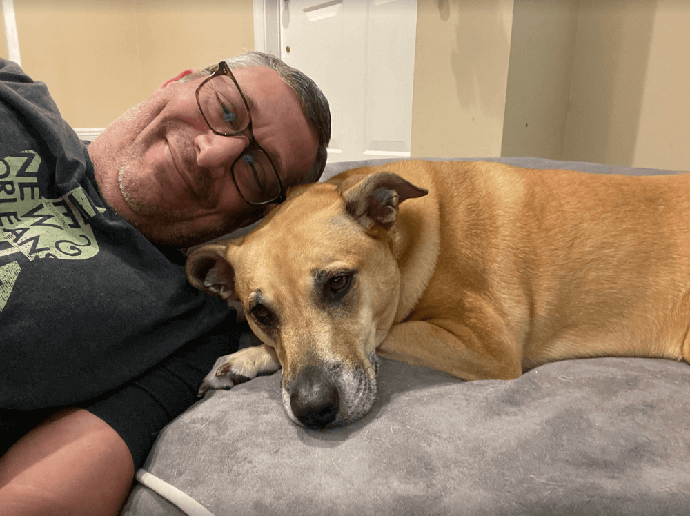 A man wearing glasses smiles whilst lying next to a large tan dog resting on a grey bed. Both appear relaxed and comfortable indoors.