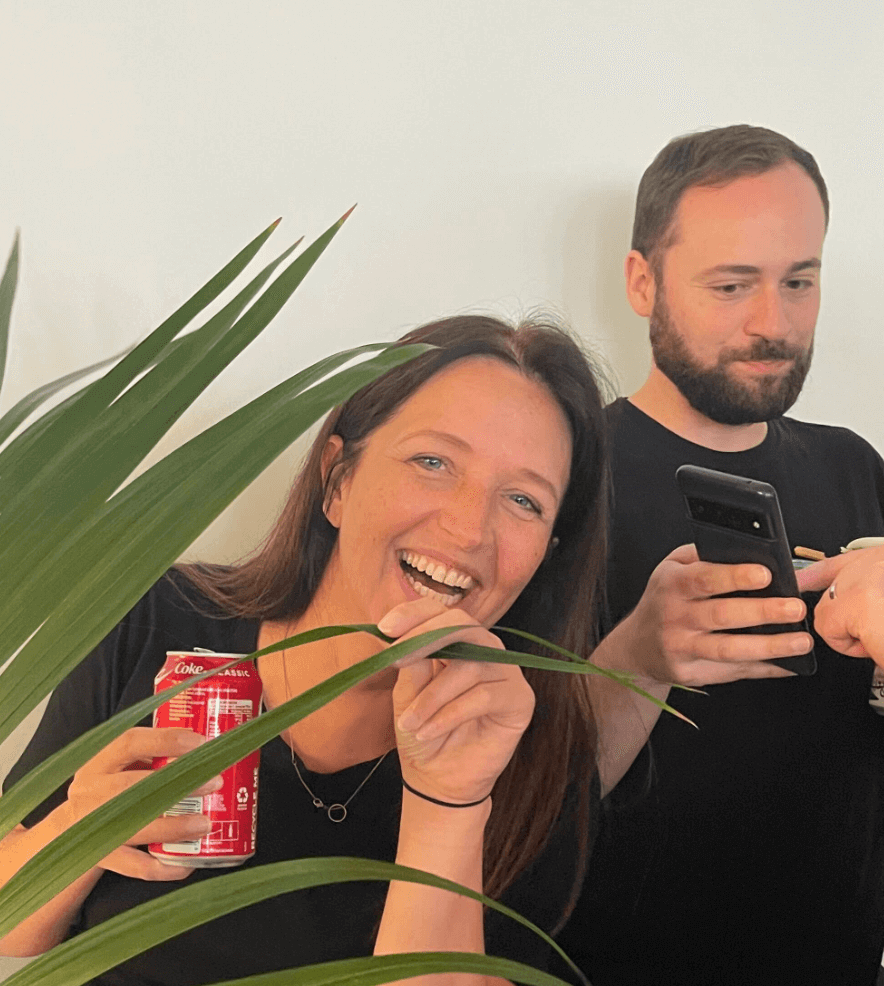 A woman smiles whilst holding a Coke can and playfully posing with a plant. A man in the background looks at his mobile. Both wear black shirts.