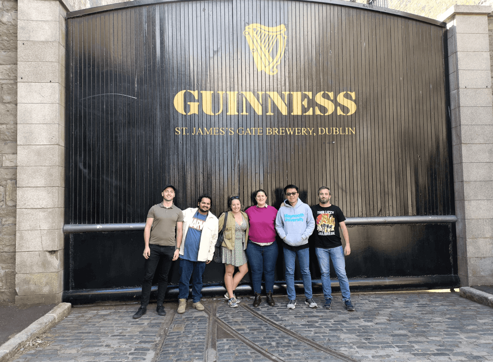 Six people stand in front of the large black gate at Guinness St. James's Gate Brewery in Dublin, featuring the brewery's gold harp logo and text. The group pose together on a cobbled street.
